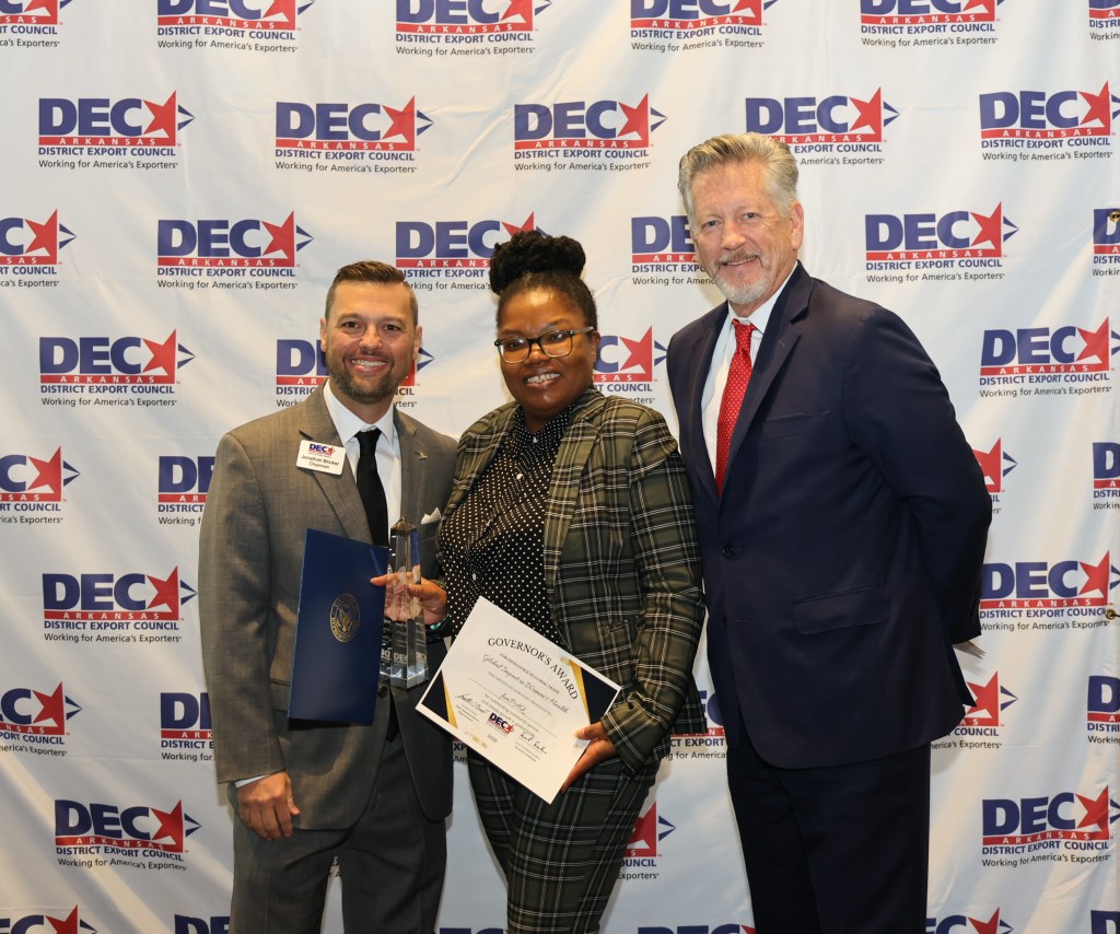 Three individuals pose for a photo at an awards ceremony, holding a Governor's Award certificate. The backdrop features the District Export Council logo.