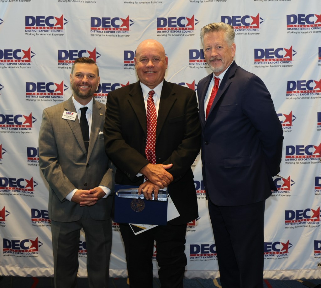 Three men in formal attire posing for a photo in front of a branded backdrop for the District Export Council.