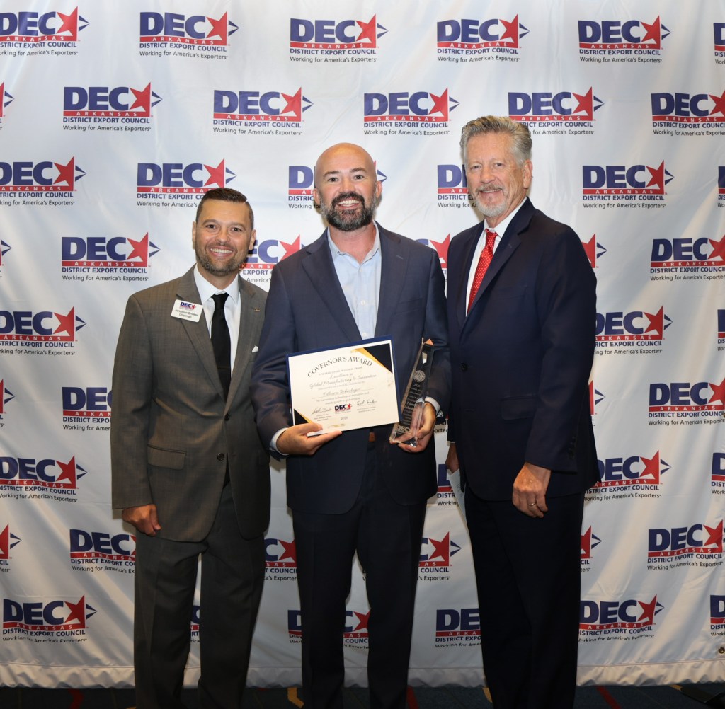 Three men stand together at an event, holding an award and certificate in front of a backdrop featuring the District Export Council logo.