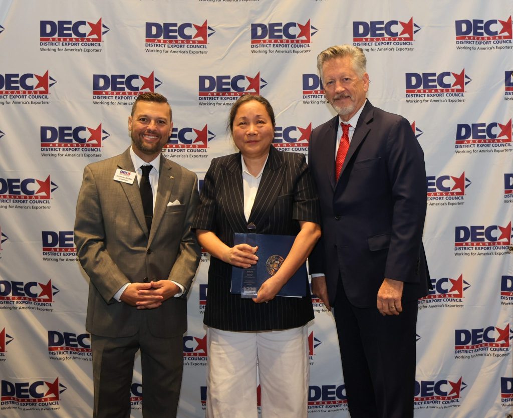 A group of three people posing for a photo in front of a banner for the District Export Council, featuring their logo. One person holds a document, while the others are smiling and dressed in formal attire.