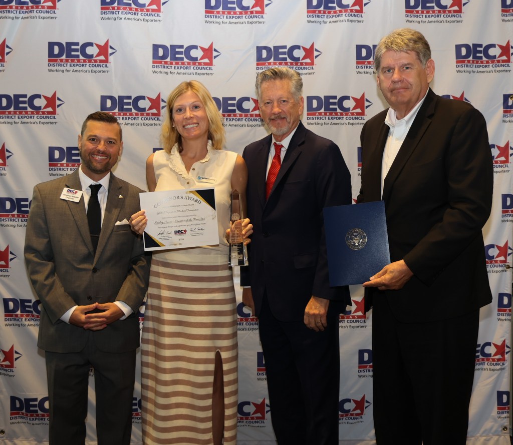 Four individuals pose at an awards ceremony, holding a certificate and a trophy, with a backdrop featuring the District Export Council logo.